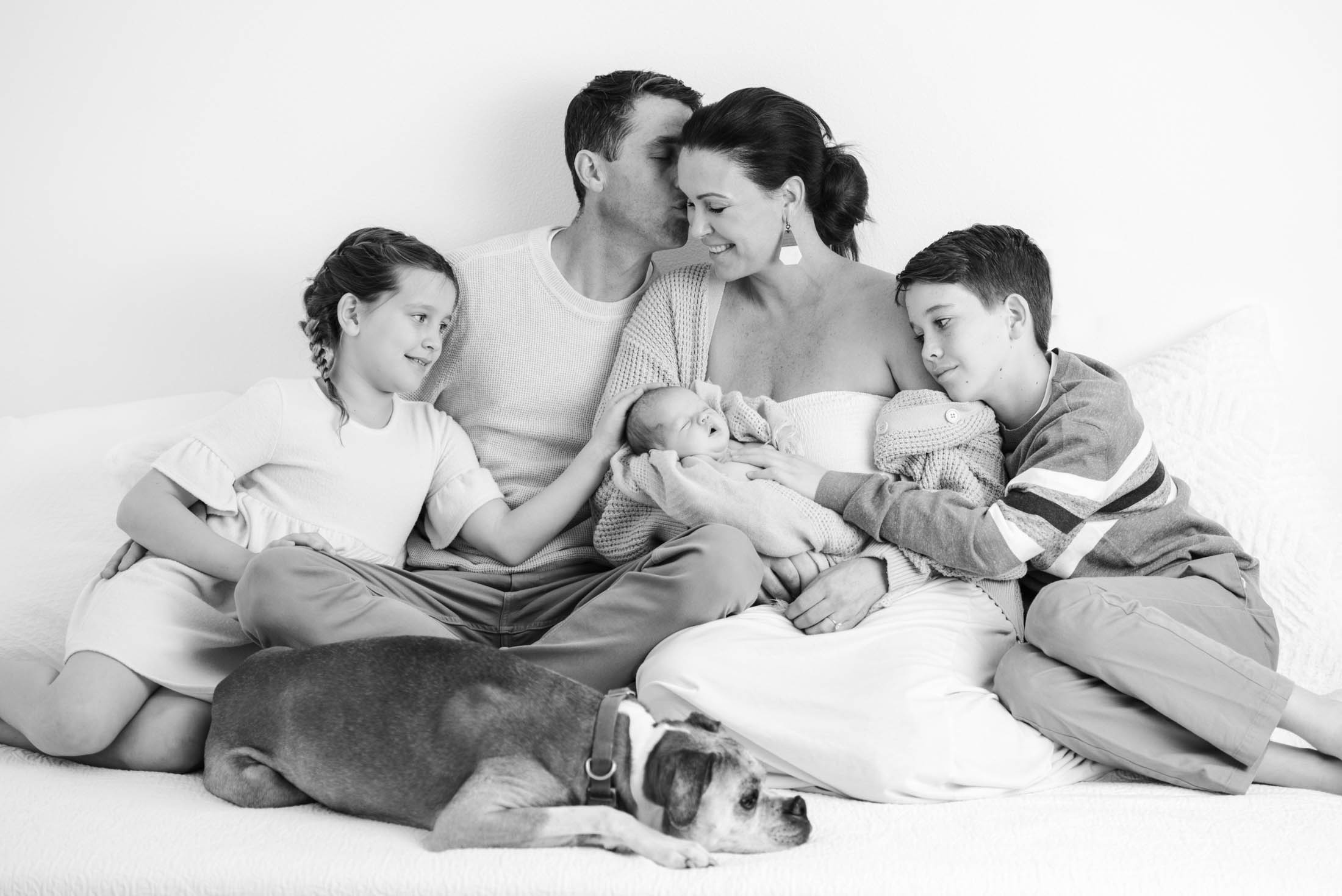A black-and-white picture showing a family of five and a dog snuggling on a bed in a studio in el dorado hills. The mother is holding a newborn baby.