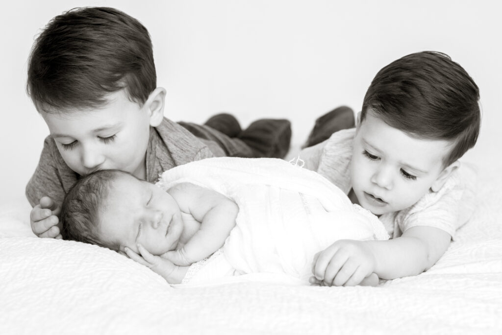 A picture of a newborn baby sleeping all wrapped in a swaddle. Behind him his older brother kisses his head and his other brother plays with his toes.