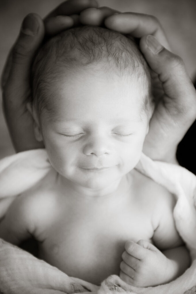 A black and white picture of a newborn baby boy held in his father's hands while wrapped loosely in a blanket. The baby is smiling.