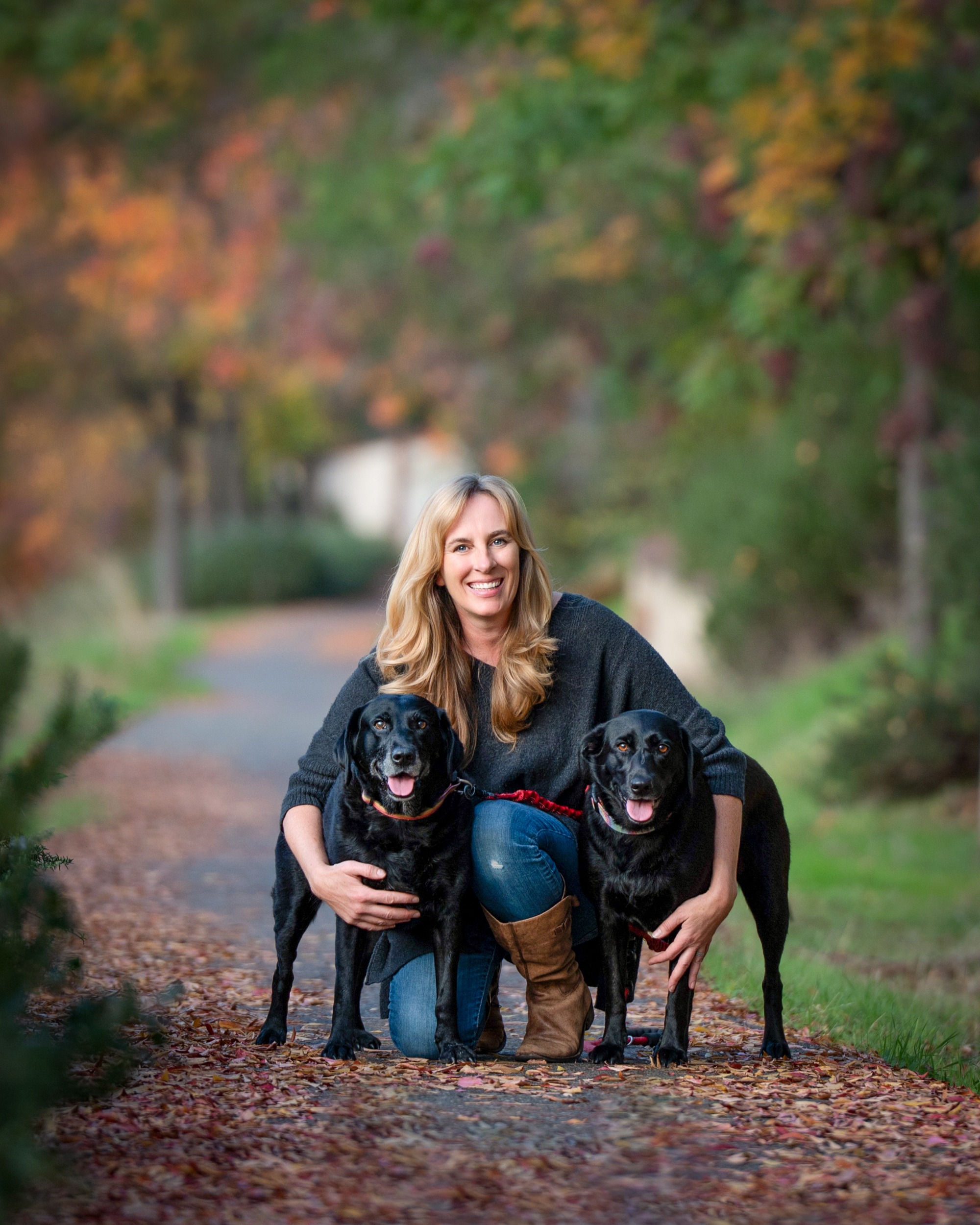 A pet photographer in El Dorado Hills hugs her 2 black labs on a fall tree lined path